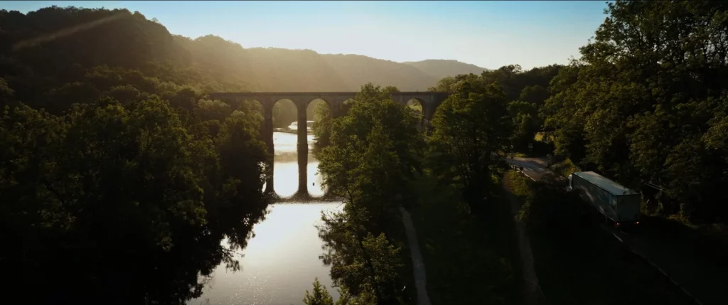Camion Le Roy Logistique traversant un paysage boisé près d’un viaduc dans la lumière du soir.