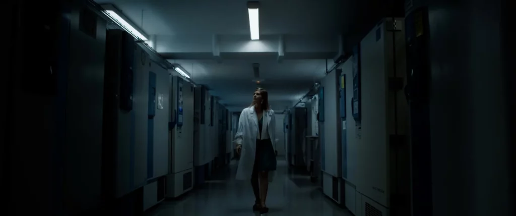 Chercheuse en blouse blanche avançant dans un couloir sombre de laboratoire.