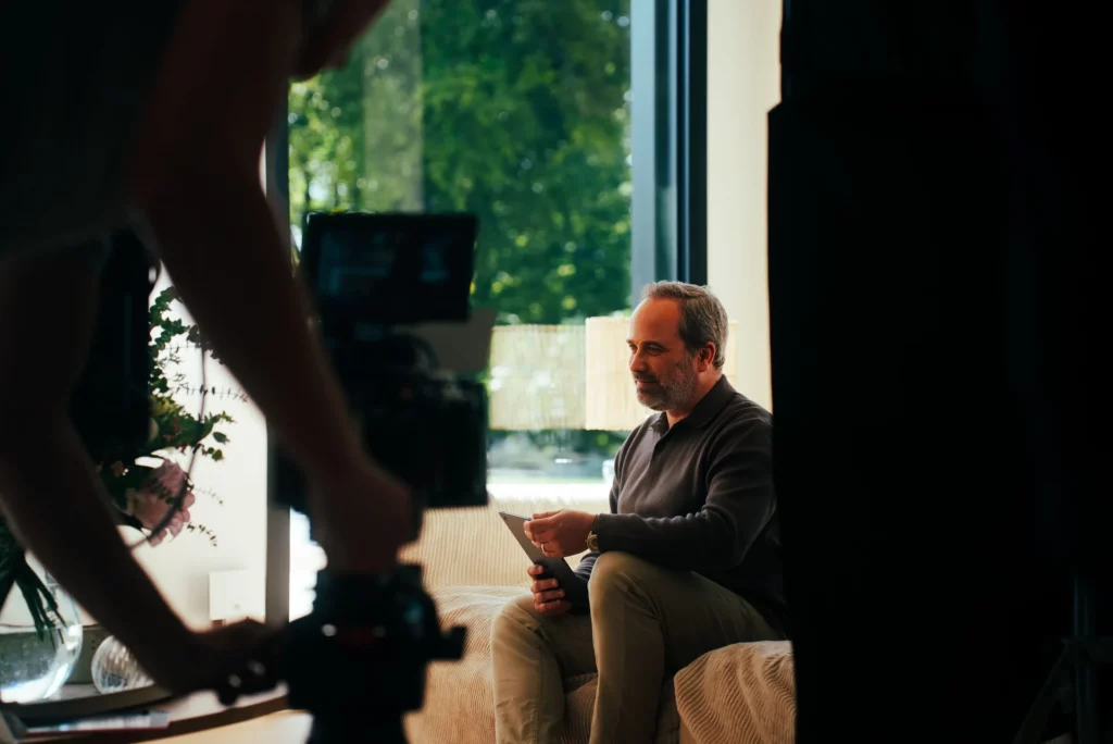 coulisses-campagne-bochassy-tournage-canape Caméra filmant un comédien assis sur le canapé dans un intérieur ouvert sur le jardin.