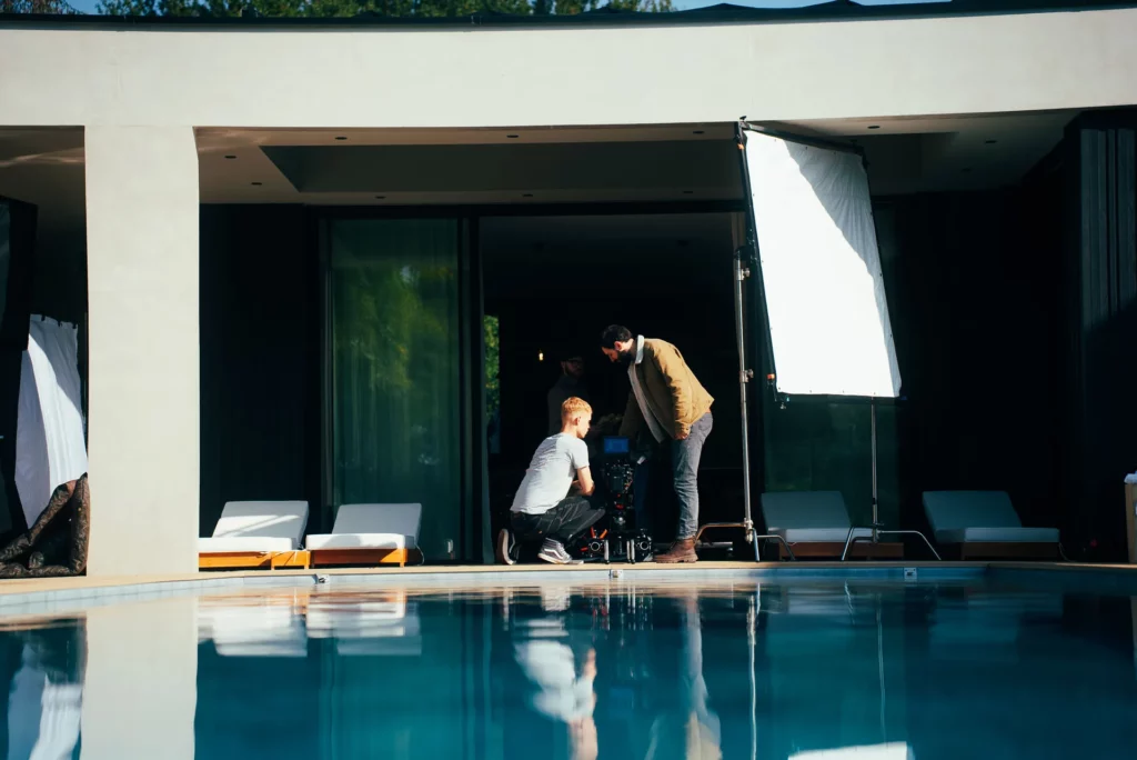 coulisses-campagne-bochassy-tournage-piscine Équipe filmant une scène près de la baie vitrée, avec un grand diffuseur installé au bord de la piscine.