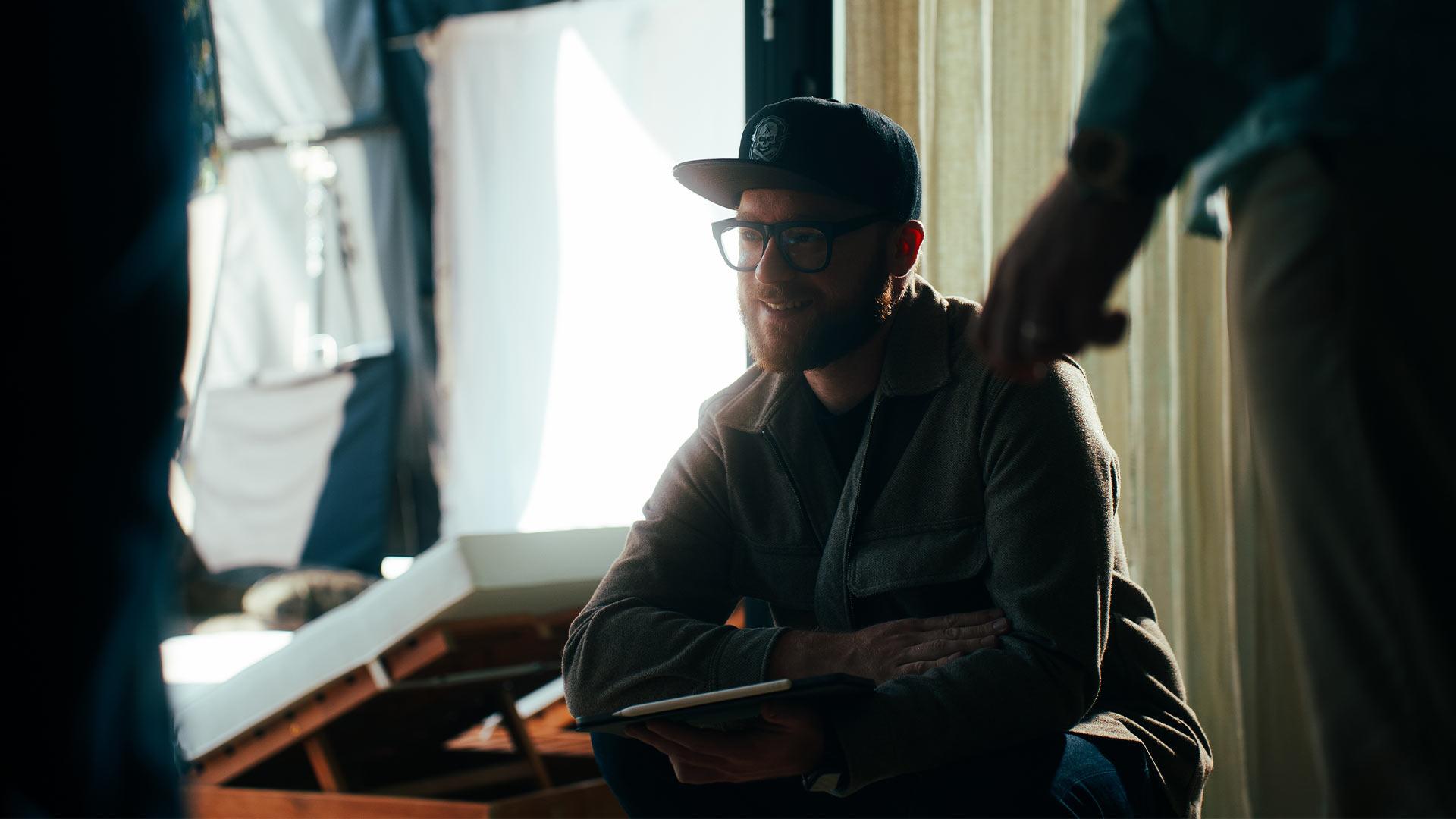 Robin Welschinger assis sur un tournage, Ipad à la main, éclairé par une lumière naturelle douce ; illustration de la gestion d’équipe, du calme et de l’expérience nécessaire sur un plateau.