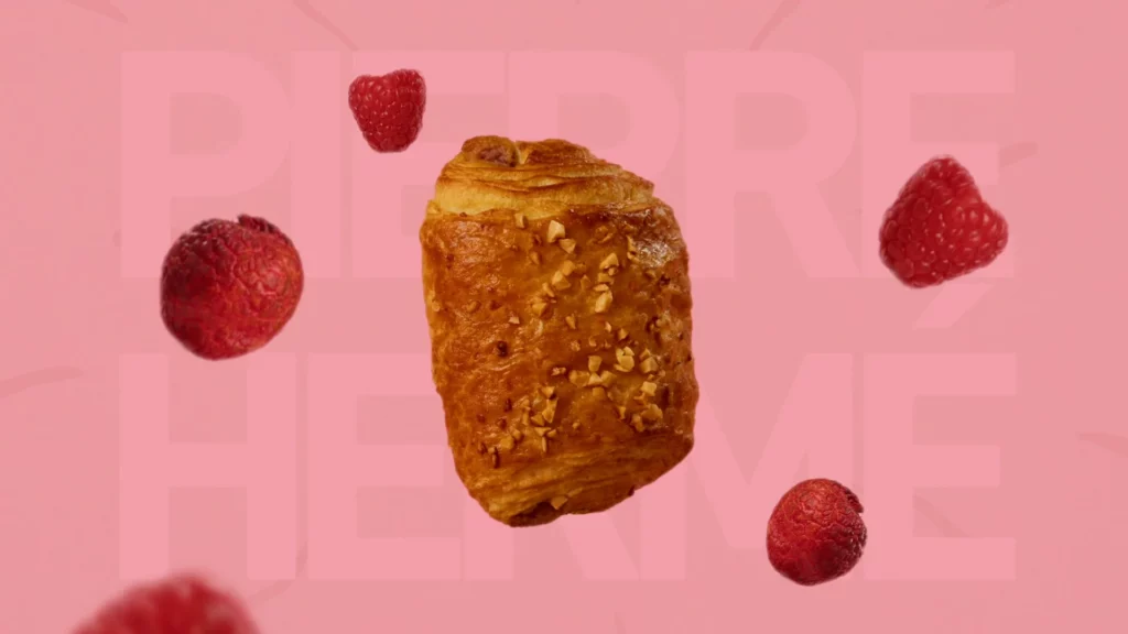 Viennoiserie Signature Framboise Litchi entourée de framboises et de litchis sur fond rose avec typographie Pierre Hermé.
