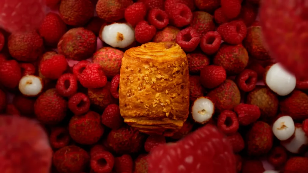 Viennoiserie Signature Framboise Litchi au centre d’une composition de framboises et de litchis sur fond rose.