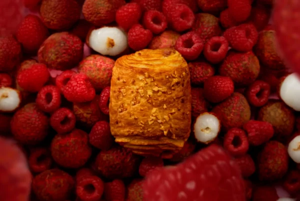 Viennoiserie Signature Framboise Litchi au centre d’une composition de framboises et de litchis sur fond rose.