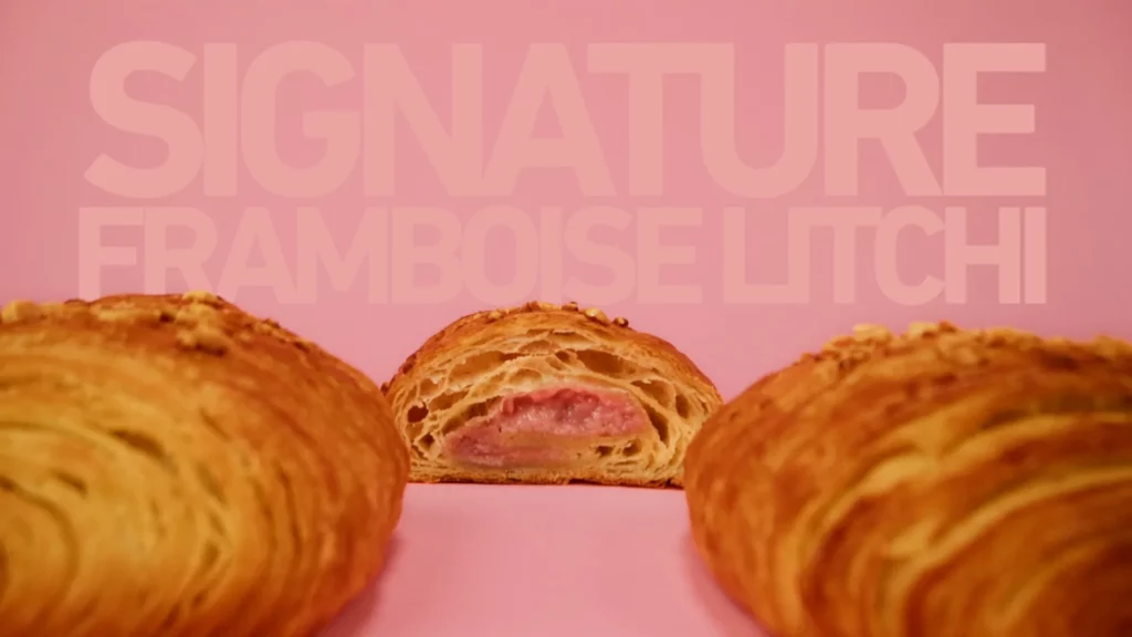 Viennoiserie Signature Framboise Litchi coupée en deux, montrant la garniture rose devant le texte Signature Framboise Litchi.