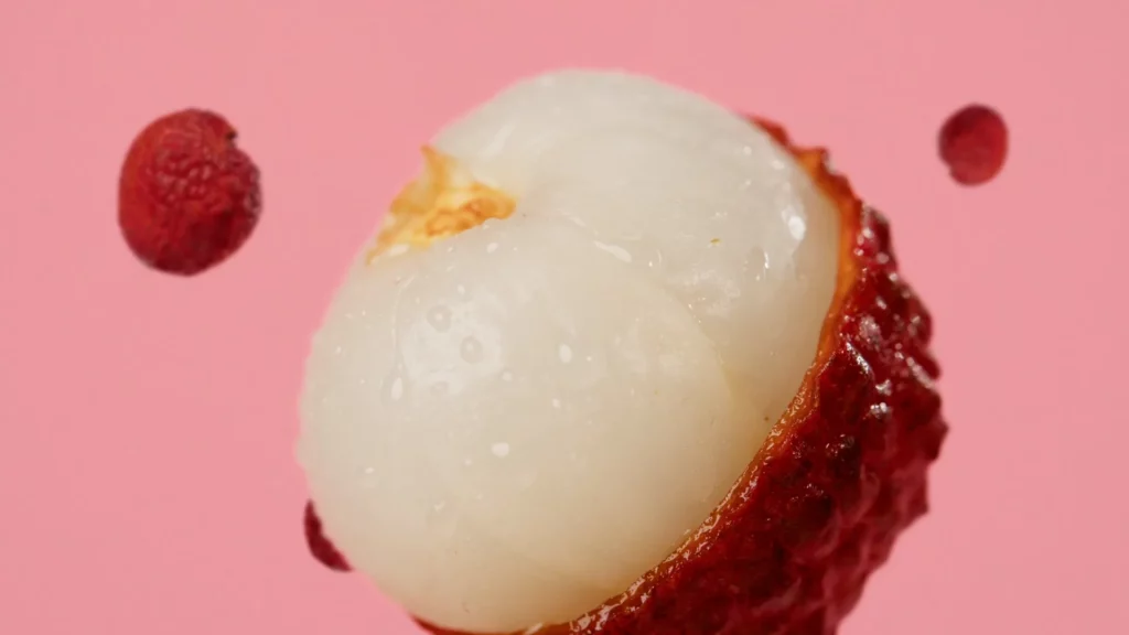 Macro d’un litchi ouvert avec chair brillante, entouré de fruits rouges sur fond rose pour le film Bridor × Pierre Hermé.