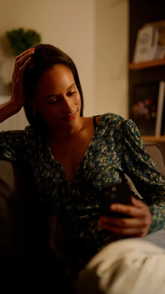 Jeune femme assise sur un canapé regardant son téléphone dans une ambiance chaleureuse et intime.