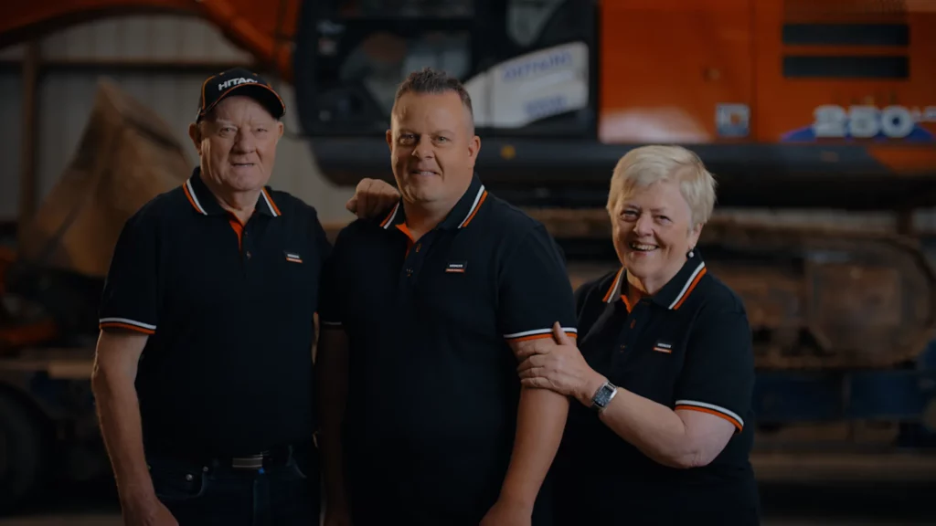 Trois membres de l’entreprise familiale Deffains TP posant devant une pelle Hitachi.
