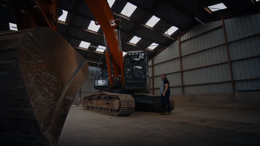 Pelle Hitachi stationnée dans un hangar, avec son godet au premier plan et un opérateur à proximité.
