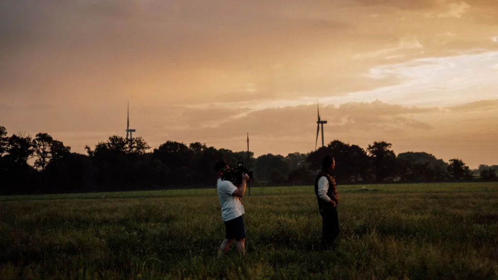 2026-ydeo-ensemble-formulons-votre-avenir-bts-champ-camera-coucher-soleil Coulisses du tournage YDEO dans un champ au coucher du soleil, avec un opérateur caméra filmant un homme.