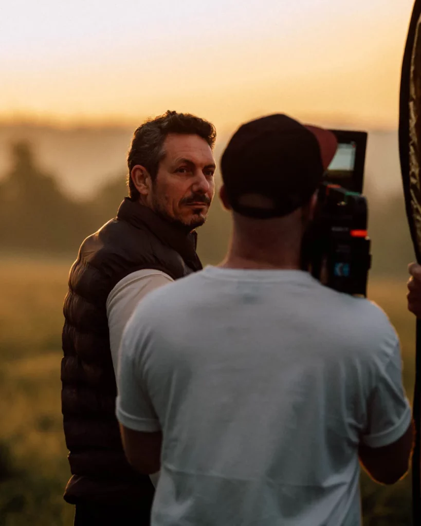 2026-ydeo-ensemble-formulons-votre-avenir-bts-champ-camera-portrait Opérateur caméra filmant un homme dans un champ au coucher du soleil pendant le tournage du film YDEO Ensemble, formulons votre avenir.