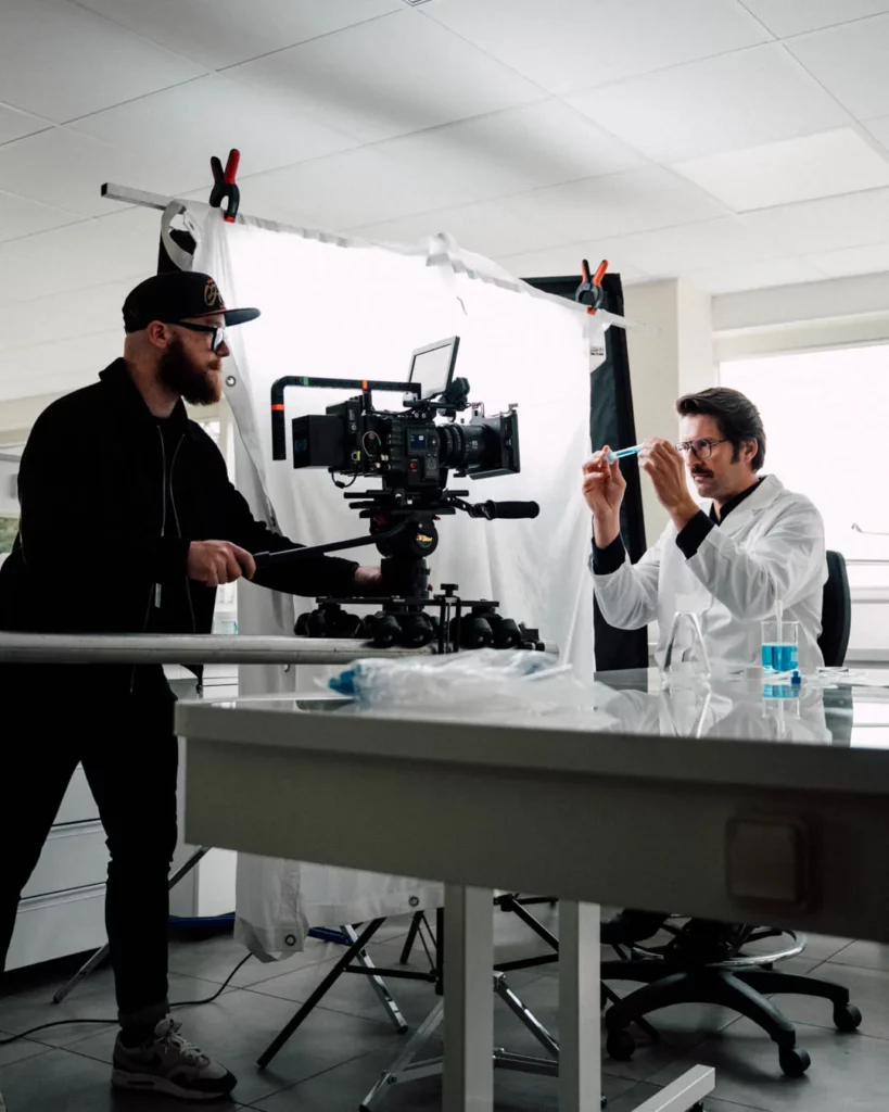 2026-ydeo-ensemble-formulons-votre-avenir-bts-laboratoire-camera-homme-blouse Coulisses du tournage YDEO en laboratoire, avec un opérateur caméra filmant un homme en blouse blanche manipulant un tube de laboratoire.