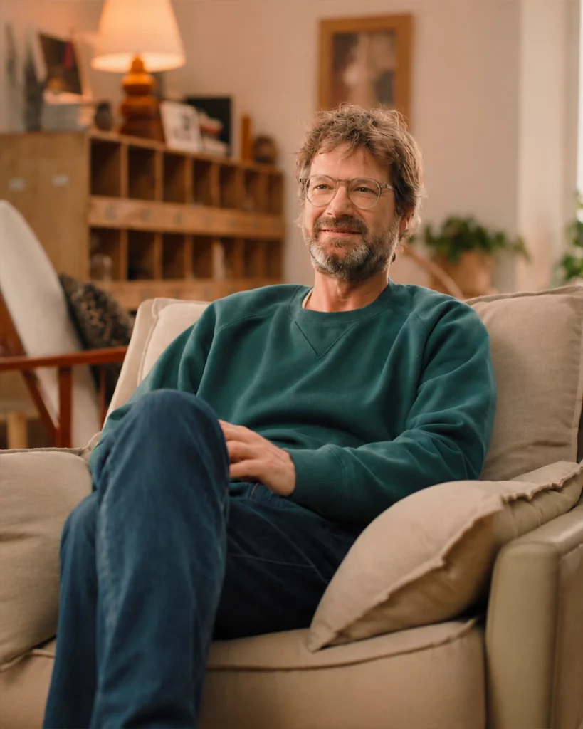 Homme assis dans un décor intérieur chaleureux, éclairé par une lumière douce.