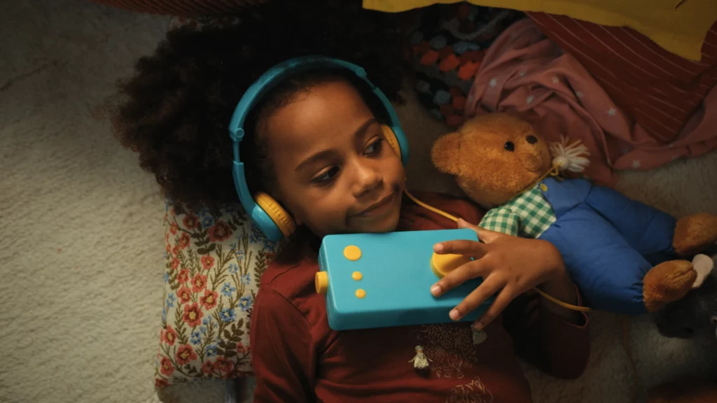 Une enfant avec un casque audio serre une boîte à histoires Lunii bleue contre elle, allongée près d'un ours en peluche.