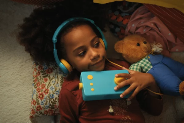 Une enfant avec un casque audio serre une boîte à histoires Lunii bleue contre elle, allongée près d'un ours en peluche.