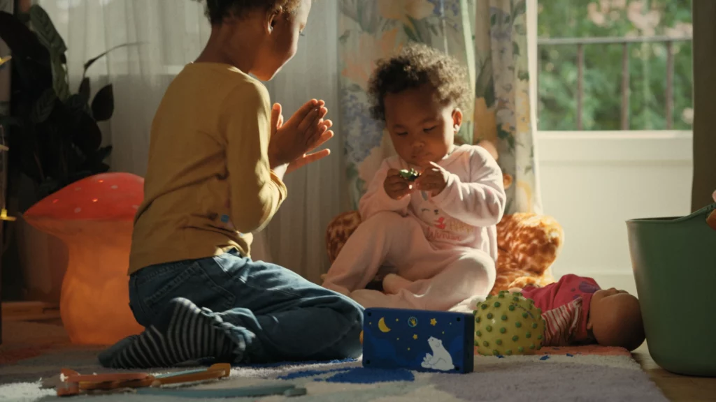 Un enfant joue avec un bébé près de la fenêtre d'une chambre, une boîte à histoires Lunii illustrée posée au premier plan.