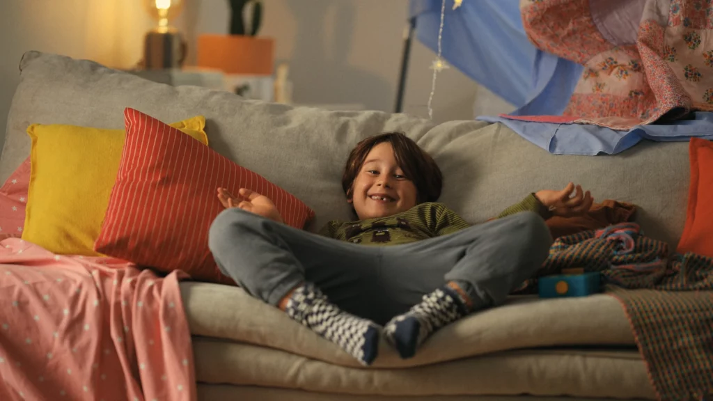 Un garçon souriant est allongé sur un canapé, entouré de coussins et de couvertures colorées dans le film Lunii.