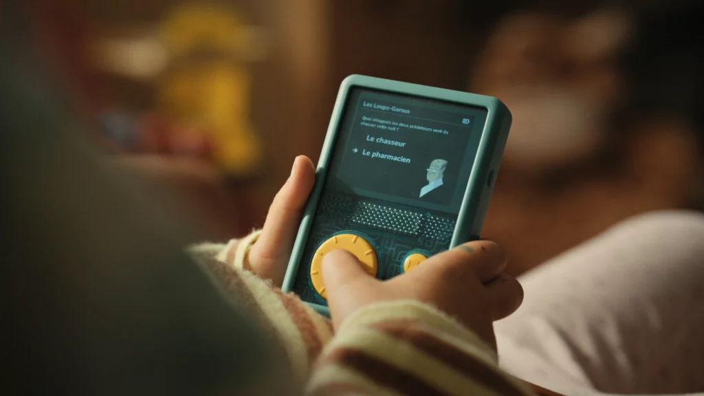 Gros plan sur les mains d'un enfant utilisant un appareil audio Lunii à écran, avec un choix de personnages affiché.