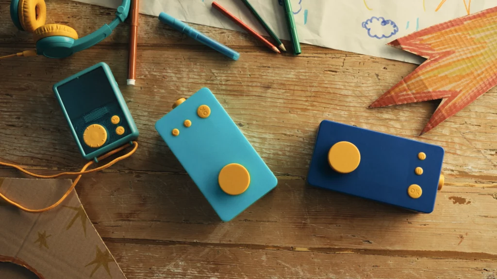 Plusieurs appareils audio et un casque pour enfants Lunii sont disposés sur une table en bois parmi des crayons et des dessins.