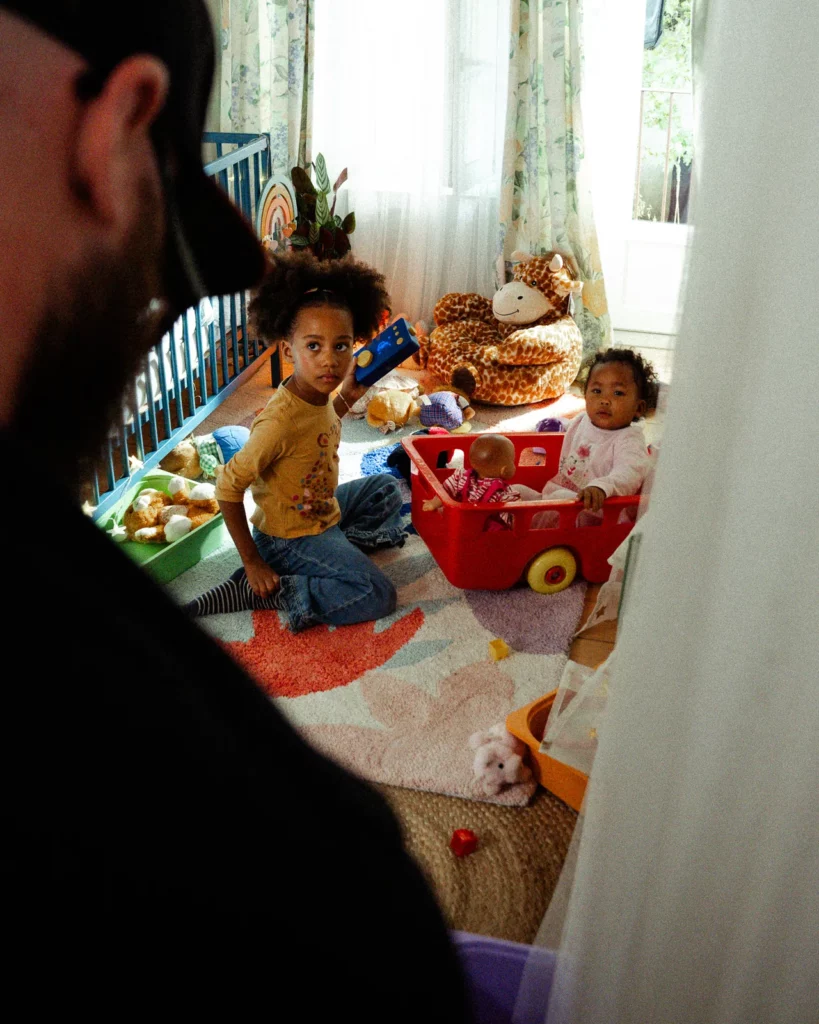 lunii-bts-enfants-decor-chambre-jouets-2026 Deux jeunes enfants attendent dans une chambre décorée de jouets pendant le tournage Lunii, avec un membre de l'équipe au premier plan.