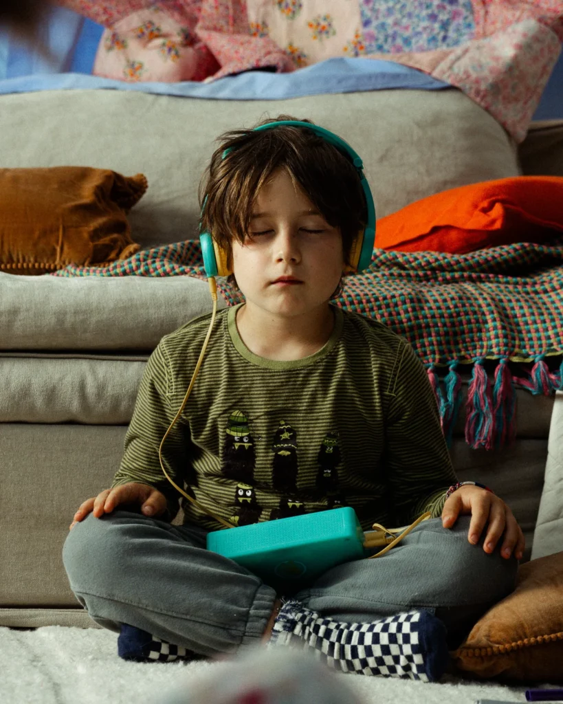 lunii-bts-garcon-casque-audio-boite-histoires-2026 Un garçon, les yeux fermés, porte un casque audio turquoise et tient une boîte à histoires Lunii pendant une pause de tournage.