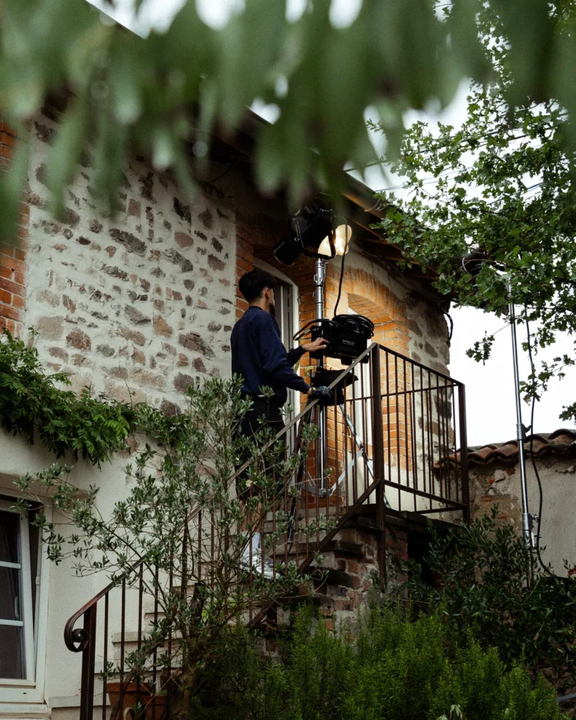 lunii-bts-installation-projecteur-escalier-exterieur-2026 Coulisses du tournage Lunii : un technicien installe un projecteur sur un escalier extérieur devant une maison en pierre.