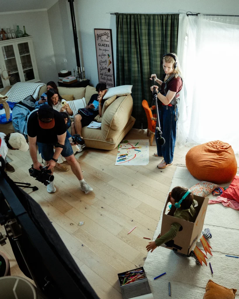lunii-bts-plateau-equipe-perche-enfant-fusee-2026 Plan large des coulisses du tournage Lunii montrant la cadreuse, la perchiste et un enfant dans une fusée en carton.