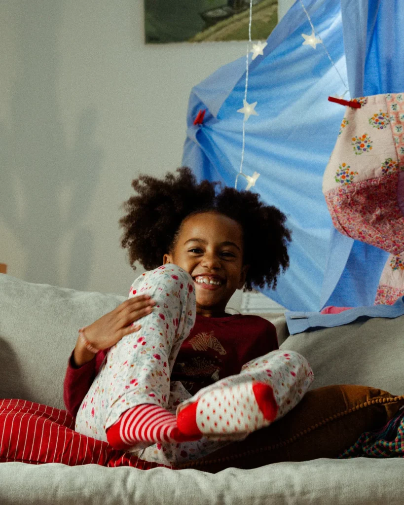 lunii-bts-portrait-enfant-souriante-canape-2026 Portrait en coulisses d'une enfant souriante, assise sur un canapé au milieu des tissus et des coussins du décor Lunii.